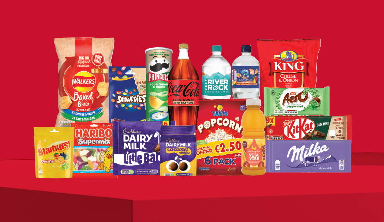 A promotional image showcasing a variety of snack foods and drinks against a red backdrop. Featured items include Walkers crisps, Pringles, Coca-Cola, River Rock water, King crisps, Aero and KitKat chocolate bars, Haribo sweets, and Milka chocolate.