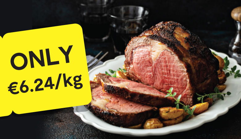 Promotional image showcasing a cooked leg of lamb garnished with roasted garlic, herbs, and mushrooms on a white platter. In the background, a side of steamed broccoli is visible. A yellow tag on the left displays the price: 'ONLY €11.25/kg' in bold black text.