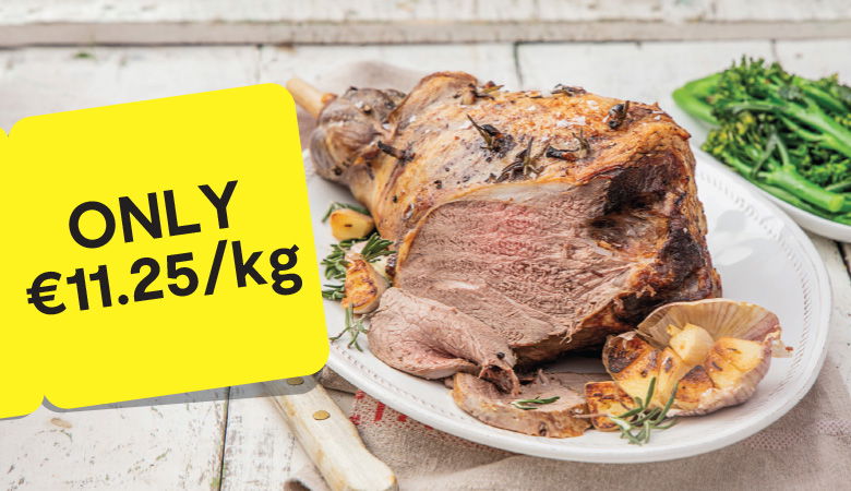 Promotional image showcasing a cooked leg of lamb garnished with roasted garlic, herbs, and mushrooms on a white platter. In the background, a side of steamed broccoli is visible. A yellow tag on the left displays the price: 'ONLY €11.25/kg' in bold black text.