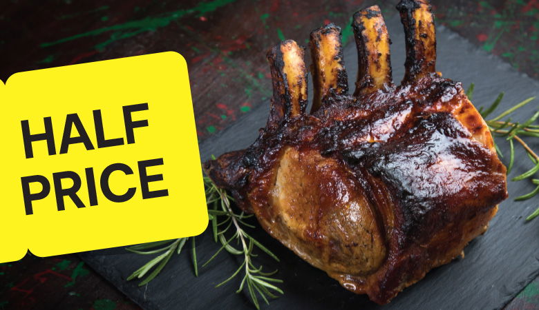 A top-down photo of a roasted pork loin sliced and served on a black plate with roasted potatoes and vegetables. The plate is resting on a wooden table. A yellow tag with the text "HALF PRICE" is placed in the top left corner.