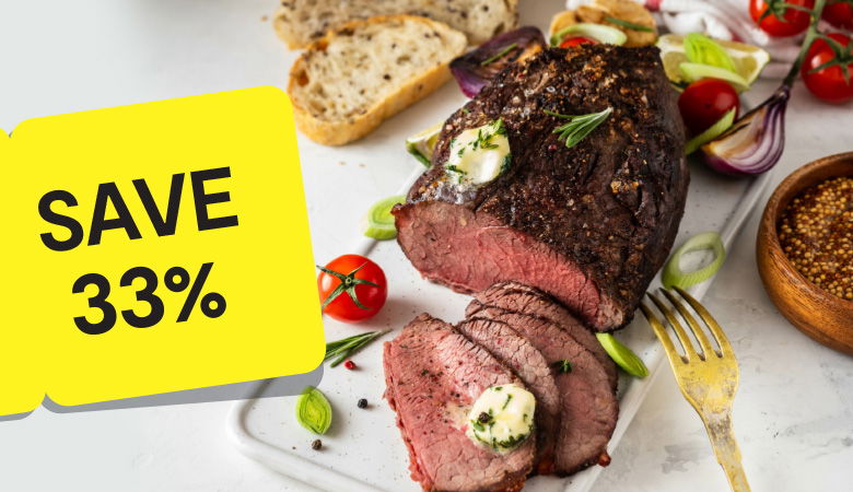 A top-down photo of a roasted beef joint with a few slices cut off. The beef is resting on a white plate with cherry tomatoes, rosemary sprigs, and sliced bread. A yellow tag in the foreground reads "SAVE 33%".