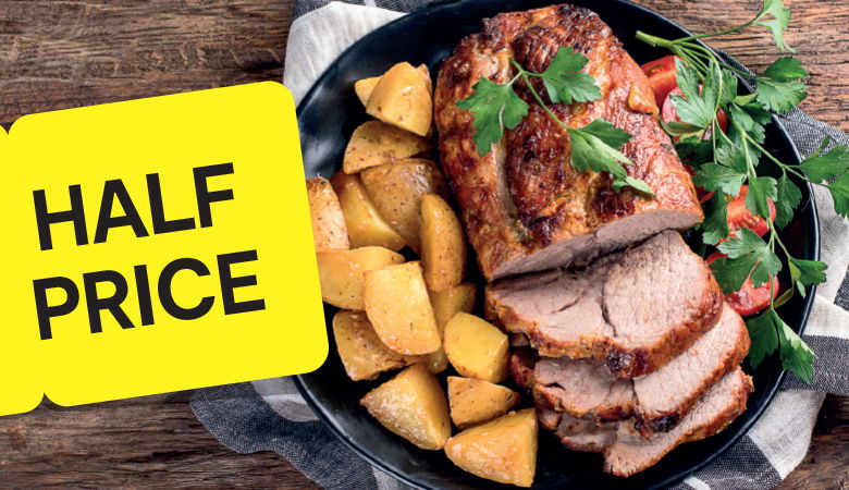 A top-down photo of a roasted pork loin sliced and served on a black plate with roasted potatoes and vegetables. The plate is resting on a wooden table. A yellow tag with the text "HALF PRICE" is placed in the top left corner.