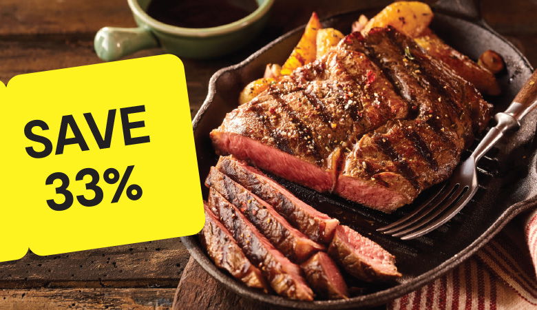 Promotional image featuring a perfectly grilled ribeye steak served in a cast iron skillet with roasted potato wedges. The steak is sliced to reveal its juicy, medium-rare interior, with a fork resting beside it. A yellow tag on the left corner prominently reads 'SAVE 33%' in bold black text, emphasizing the discount offer. In the background, a green mug of sauce adds a finishing touch to the rustic wooden table setting.