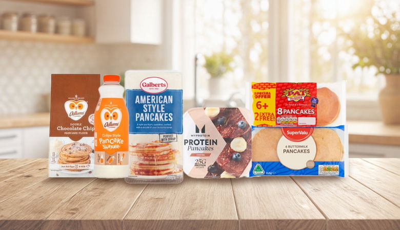 Several different pancake products are displayed on a rustic wooden table, including boxes of mix, bottles of syrup, and packages of pre-made pancakes.