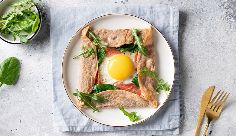 A savory crepe with a baked egg in the center, served with a fresh salad of mixed greens, cherry tomatoes, and microgreens on a white plate.