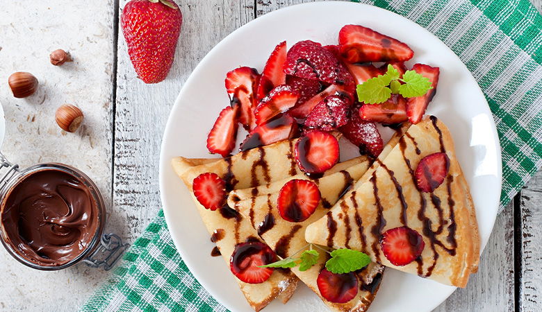 A plated crepe drizzled with chocolate sauce, topped with fresh strawberries and syrup, served with whipped cream garnished with mint and sprinkles, dusted with powdered sugar.