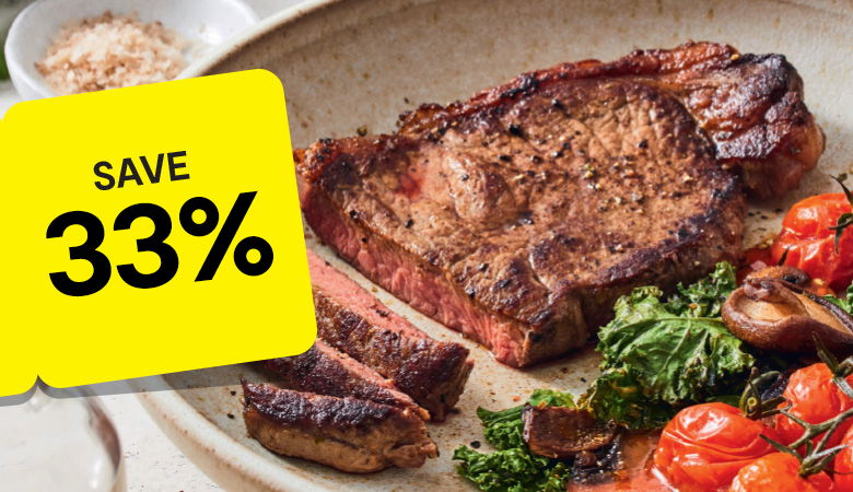 Promotional image featuring a perfectly grilled ribeye steak served in a cast iron skillet with roasted potato wedges. The steak is sliced to reveal its juicy, medium-rare interior, with a fork resting beside it. A yellow tag on the left corner prominently reads 'SAVE 33%' in bold black text, emphasizing the discount offer. In the background, a green mug of sauce adds a finishing touch to the rustic wooden table setting.