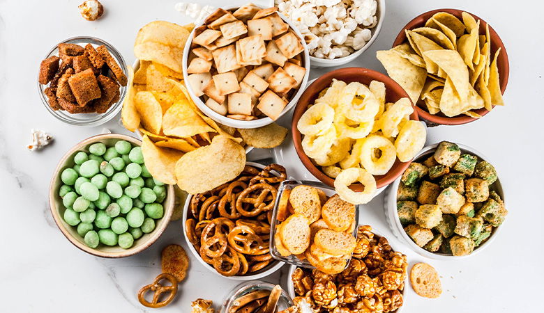 An assortment of savory and crunchy snacks in bowls, including pretzels, popcorn, potato chips, crackers, caramel popcorn, tortilla chips, and green-coated nuts, arranged on a white surface.