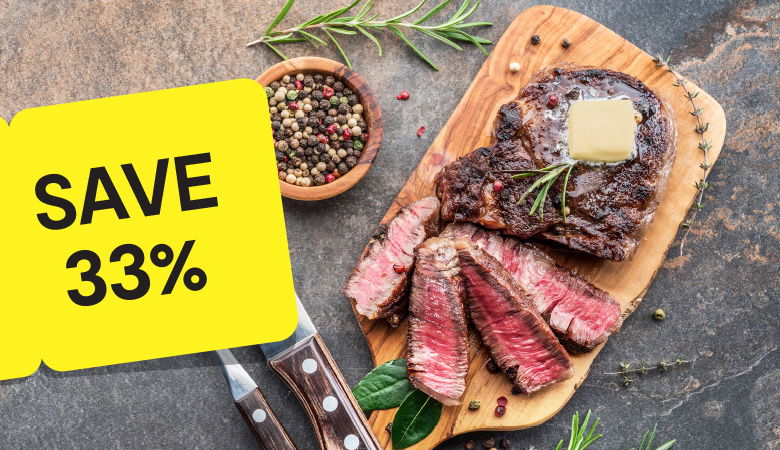 Promotional image featuring a perfectly grilled ribeye steak served in a cast iron skillet with roasted potato wedges. The steak is sliced to reveal its juicy, medium-rare interior, with a fork resting beside it. A yellow tag on the left corner prominently reads 'SAVE 33%' in bold black text, emphasizing the discount offer. In the background, a green mug of sauce adds a finishing touch to the rustic wooden table setting.