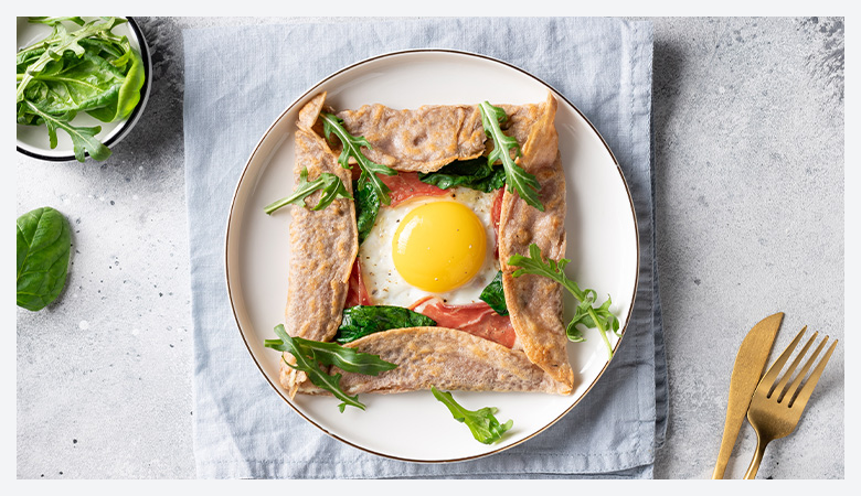 A savory crepe with a baked egg in the center, served with a fresh salad of mixed greens, cherry tomatoes, and microgreens on a white plate.