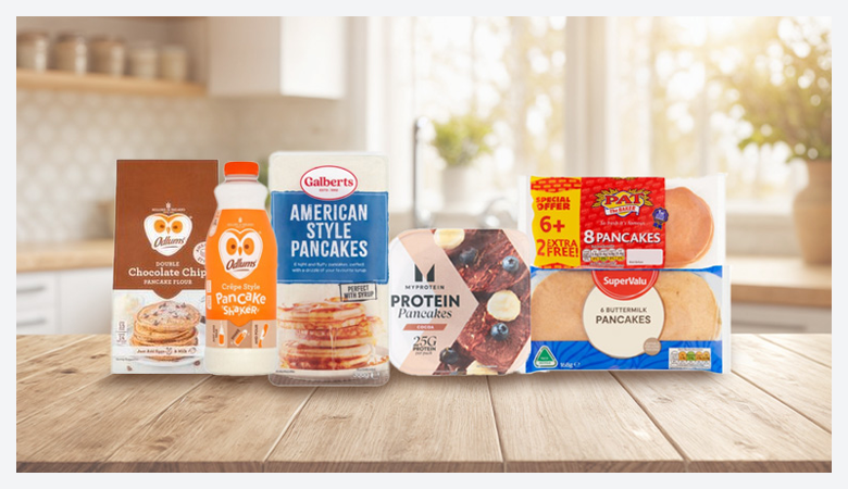 Several different pancake products are displayed on a rustic wooden table, including boxes of mix, bottles of syrup, and packages of pre-made pancakes.