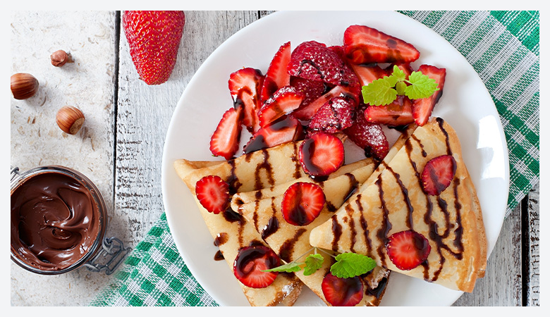 A plated crepe drizzled with chocolate sauce, topped with fresh strawberries and syrup, served with whipped cream garnished with mint and sprinkles, dusted with powdered sugar.