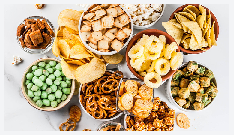 An assortment of savory and crunchy snacks in bowls, including pretzels, popcorn, potato chips, crackers, caramel popcorn, tortilla chips, and green-coated nuts, arranged on a white surface.