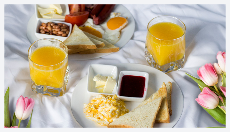 Two breakfast plates with toast, scrambled eggs, butter, jam, baked beans, sausages, fried eggs, and orange juice, placed on a white bedsheet with pink tulips.