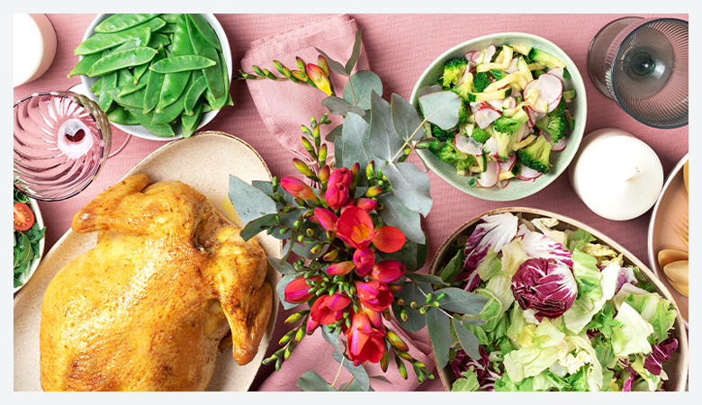 A roasted whole chicken served on a platter, accompanied by fresh salads, snap peas, and candles on a pink table setting with red flowers.