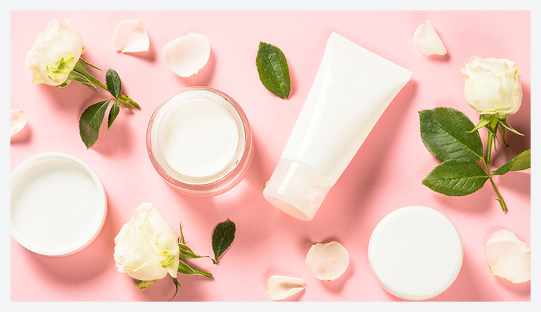 Soft, feminine beauty product arrangement with white roses and cream on a light pink background.